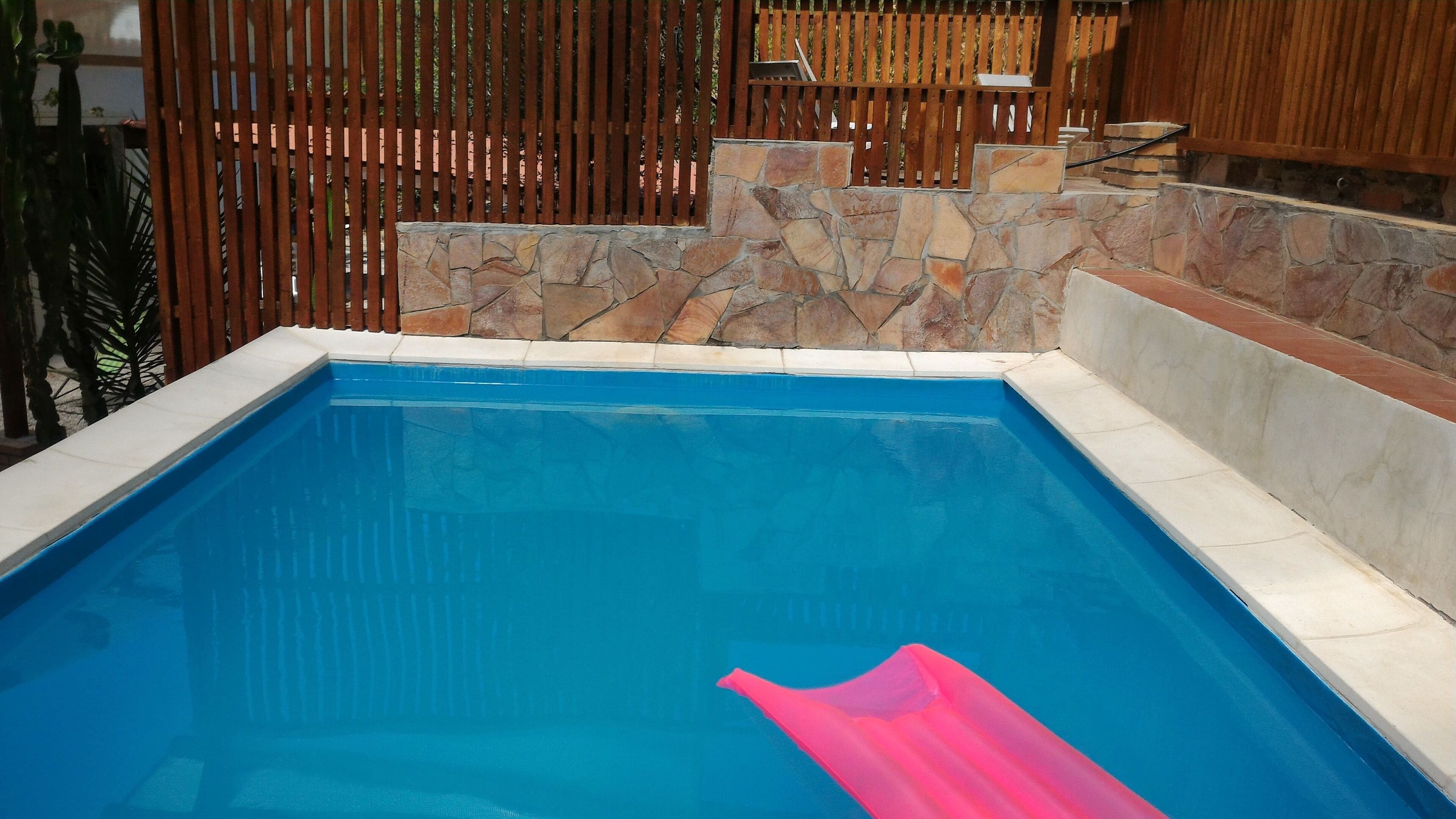 A rectangular swimming pool with a stone wall and wooden fence.