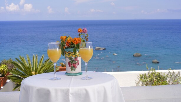 A table with two glasses of orange juice and a floral vase, set against a backdrop of the sea and sky. - Room in B&B - Ischia, 1 Relaxing Doubles Hotel Imperamare (Forio)