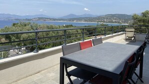 Une terrasse extérieure avec une table et des chaises, offrant une vue sur un paysage côtier avec des montagnes et un ciel dégagé.