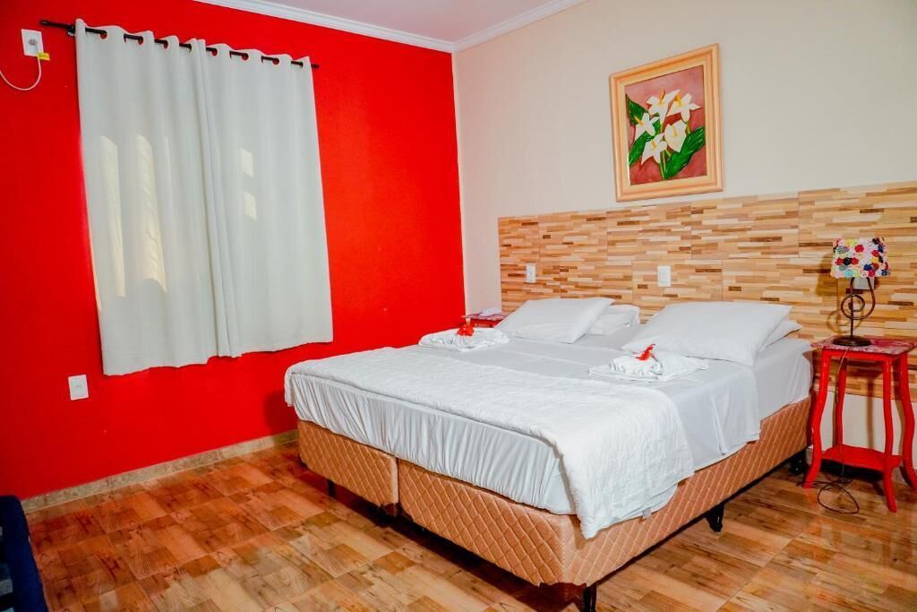 A hotel room with a red wall, a wooden headboard, a bed with white bedding, a small red stool, and a painting on the wall.