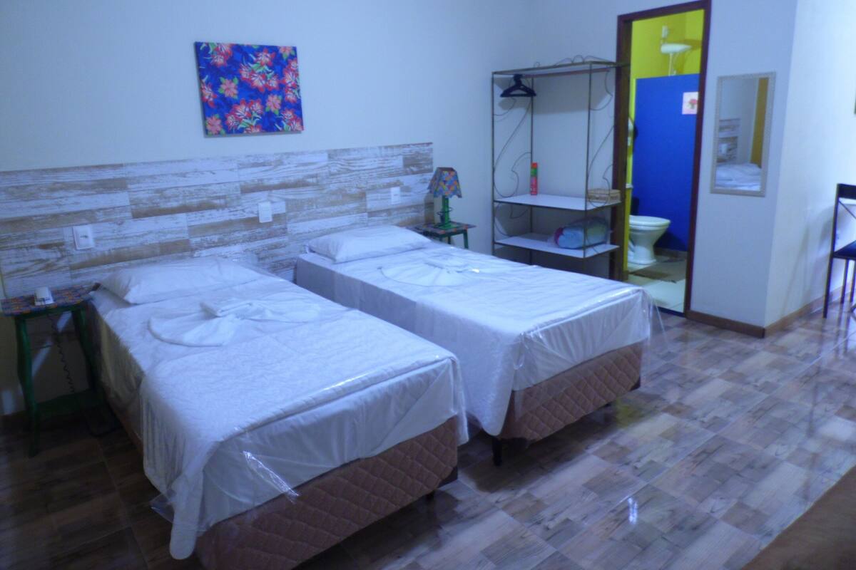 A hotel room with two beds, a wooden headboard, a colorful painting, a small table with a lamp, a shelf with toiletries, and a mirror.