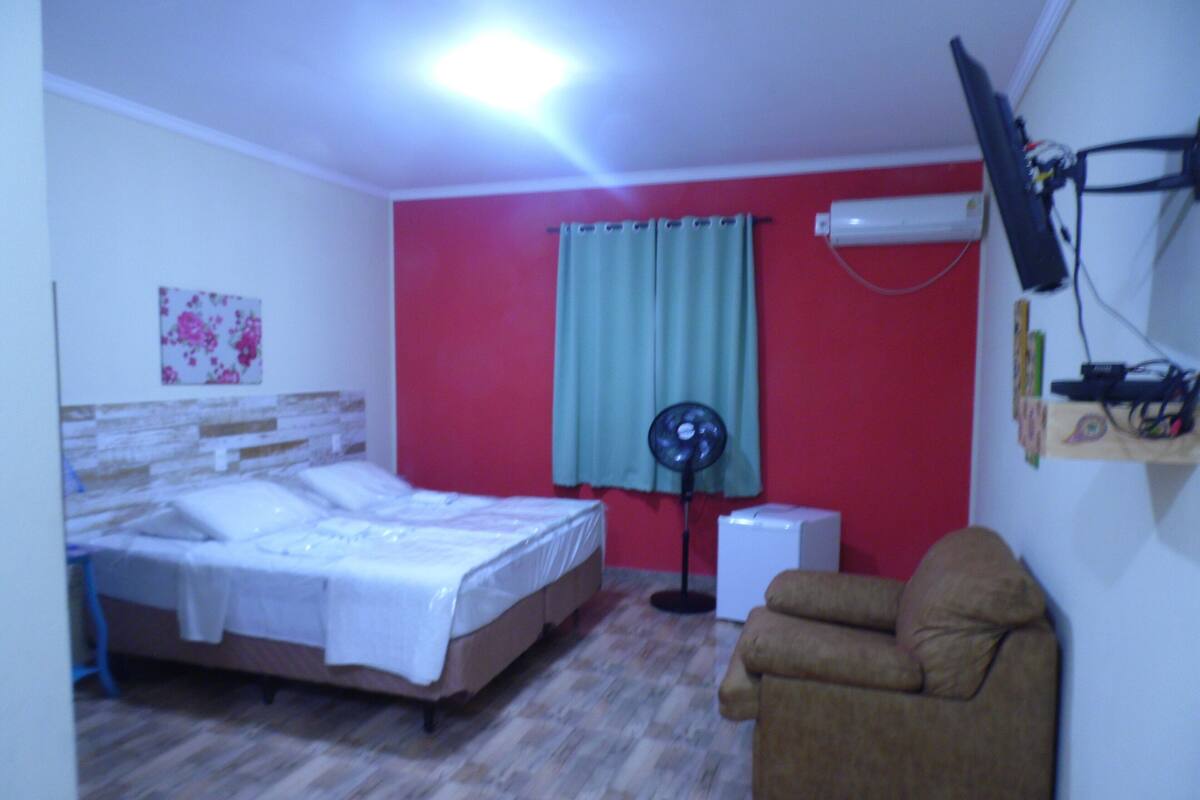 A bedroom with a bed, a chair, a standing fan, and a wall-mounted air conditioner.