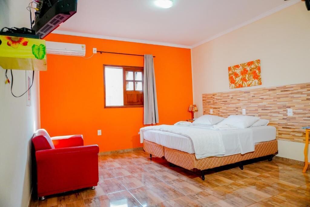 Una habitación con una cama, una silla roja, un equipo de aire acondicionado y una ventana con cortinas.