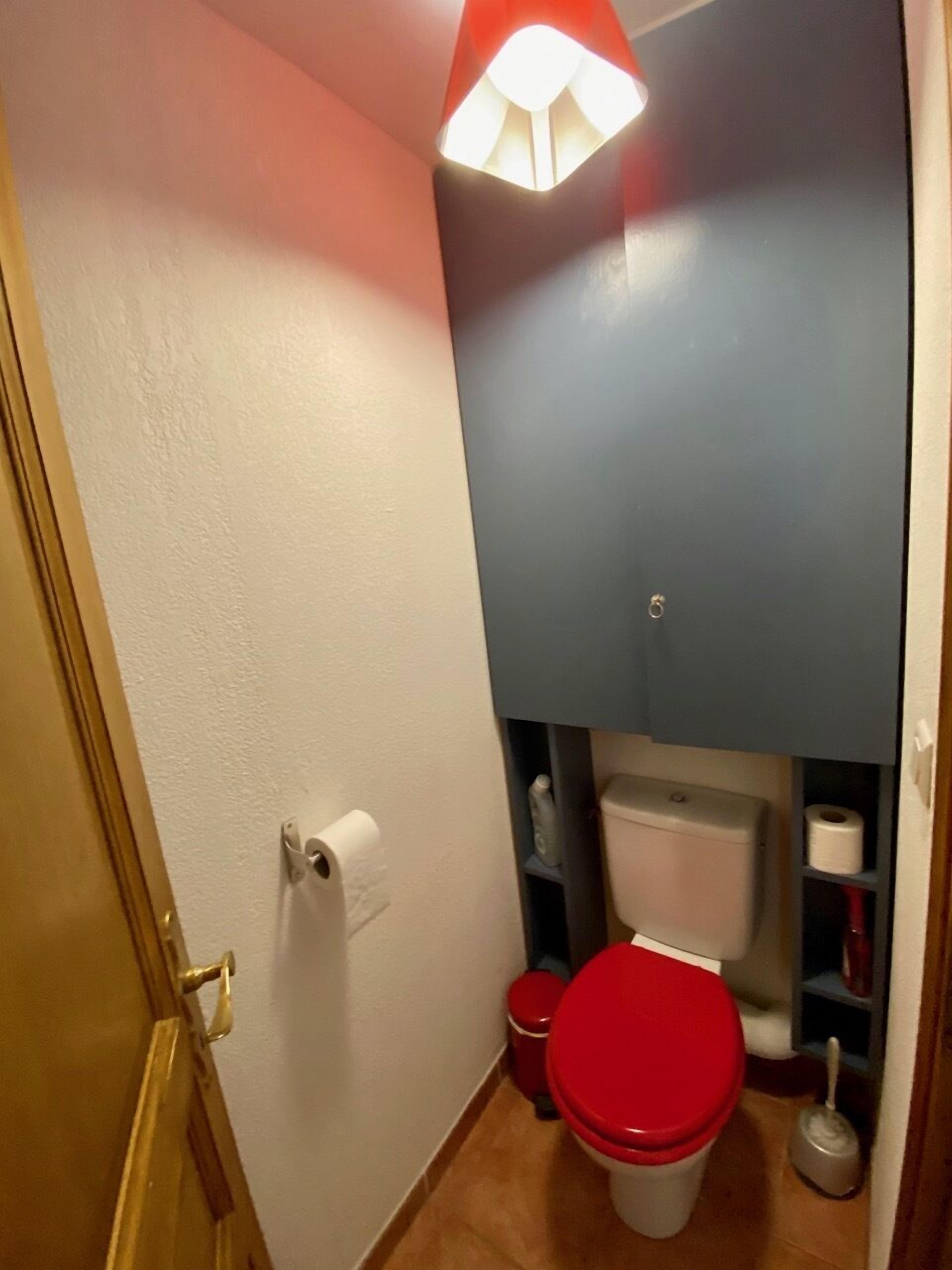 Un baño pequeño con inodoro, un asiento de inodoro rojo, un dispensador de papel higiénico blanco y un cubo de basura rojo.