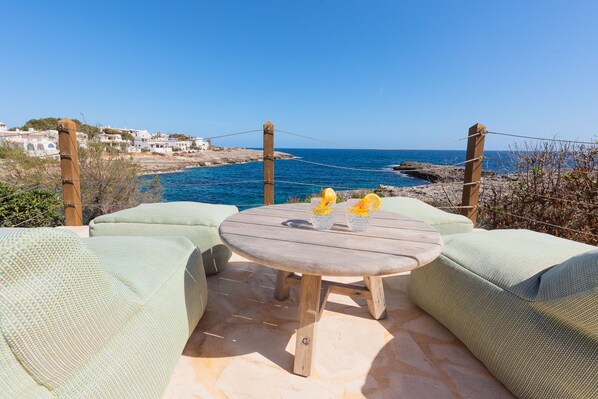 A wooden table with two yellow vases, surrounded by green cushioned seating, overlooking a rocky coastline with clear blue sea and white buildings in the background.