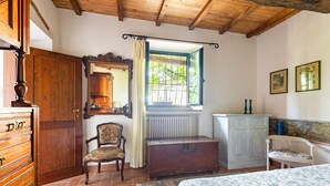 Ein Schlafzimmer mit einem Bett, einem Sessel, einem Schminktisch und einem Spiegel, mit Blick auf Grünflächen.