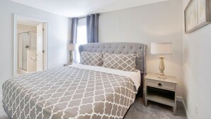 Une chambre à coucher avec un lit, une table de chevet avec une lampe, une porte donnant sur une salle de bain et une fenêtre avec des rideaux.
