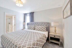 Un dormitorio con cama, mesita de noche con lámpara, puerta que da al baño y ventana con cortinas.