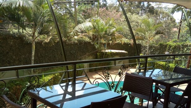 Un espacio de estar al aire libre con techo, con mesa de vidrio y sillas de mimbre, con vista a una piscina y abundante vegetación.