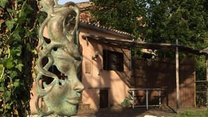 Un espacio exterior rústico con una escultura de metal, una mesa de madera y un edificio con techo de tejas.