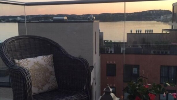 A balcony with a wicker chair and a potted plant, overlooking a body of water and buildings. - StayPlus Ocean View Apartment Balcony (Kristiansand)
