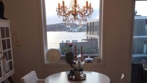 A dining area with a round table and white chairs, a chandelier, and a view of a body of water through a large window. - StayPlus Ocean View Apartment Balcony (Kristiansand)