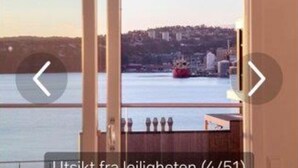 A view from a room with a large window overlooking a body of water and a distant cityscape. - StayPlus Ocean View Apartment Balcony (Kristiansand)
