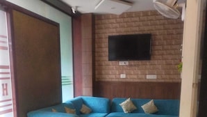 A modern living area with a blue sectional sofa, a flat-screen TV mounted on a brick wall, and a ceiling fan. - Hotel Janki Inn Varanasi (Varanasi)