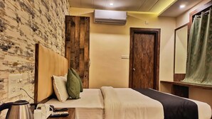 A hotel room with a wooden headboard, a bed with white and black bedding, a bedside table with a kettle and remote control, and a wooden floor. - Hotel Janki Inn Varanasi (Varanasi)