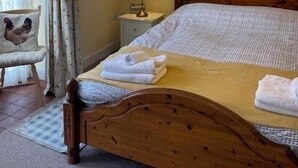 Ein ordentlich bezogenes Bett mit Holz-Kopfstück, ein Nachttisch mit Lampe und ein Sessel mit Kissen in Form eines Hahns.