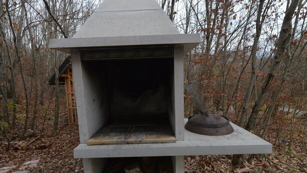 A concrete outdoor fireplace with a metal lid, surrounded by fallen leaves and trees. - Chalet Katarina (Lovinac)
