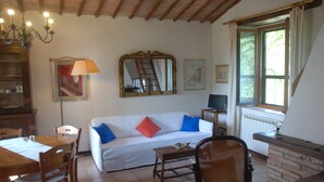 A cozy living room with a white sofa, wooden dining table, and chairs. There is a television, a lamp, and a window with curtains. - Silence and Relaxation in Countryside of Umbria (Amelia)