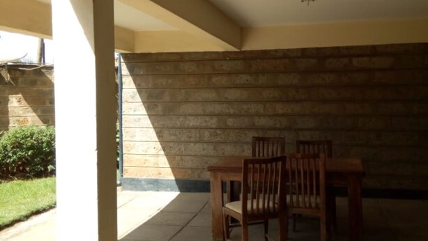 A covered patio area with a stone wall, a wooden table and chairs, and a view of a garden. - South C Bungalow (Nairobi)
