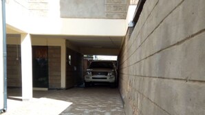 A covered parking area with a vehicle parked inside, a paved walkway, and a building with a window and a door. - South C Bungalow (Nairobi)