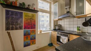 A kitchen with a stove, oven, and cabinets. There is a window with a view of buildings outside. - StayPlus Comfy 1BR Apt - Hip Area (Oslo)