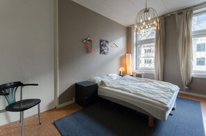 A bedroom with a bed, a chair, a small table, a lamp, and a window with curtains. - StayPlus Comfy 1BR Apt - Hip Area (Oslo)