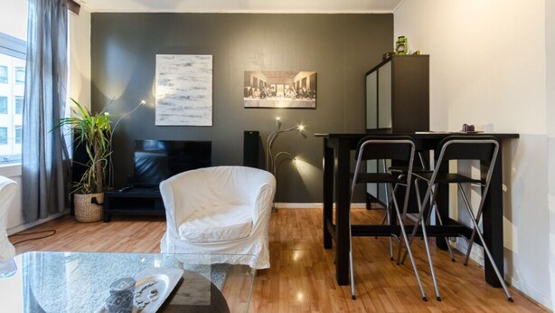 A modern living room with a sofa, a glass coffee table, a dining area with a table and chairs, a large painting, and a potted plant. - StayPlus Comfy 1BR Apt - Hip Area (Oslo)