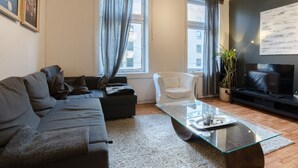 A modern living room with a grey sofa, a white armchair, a glass coffee table, and a flat-screen TV on a black stand. - StayPlus Comfy 1BR Apt - Hip Area (Oslo)