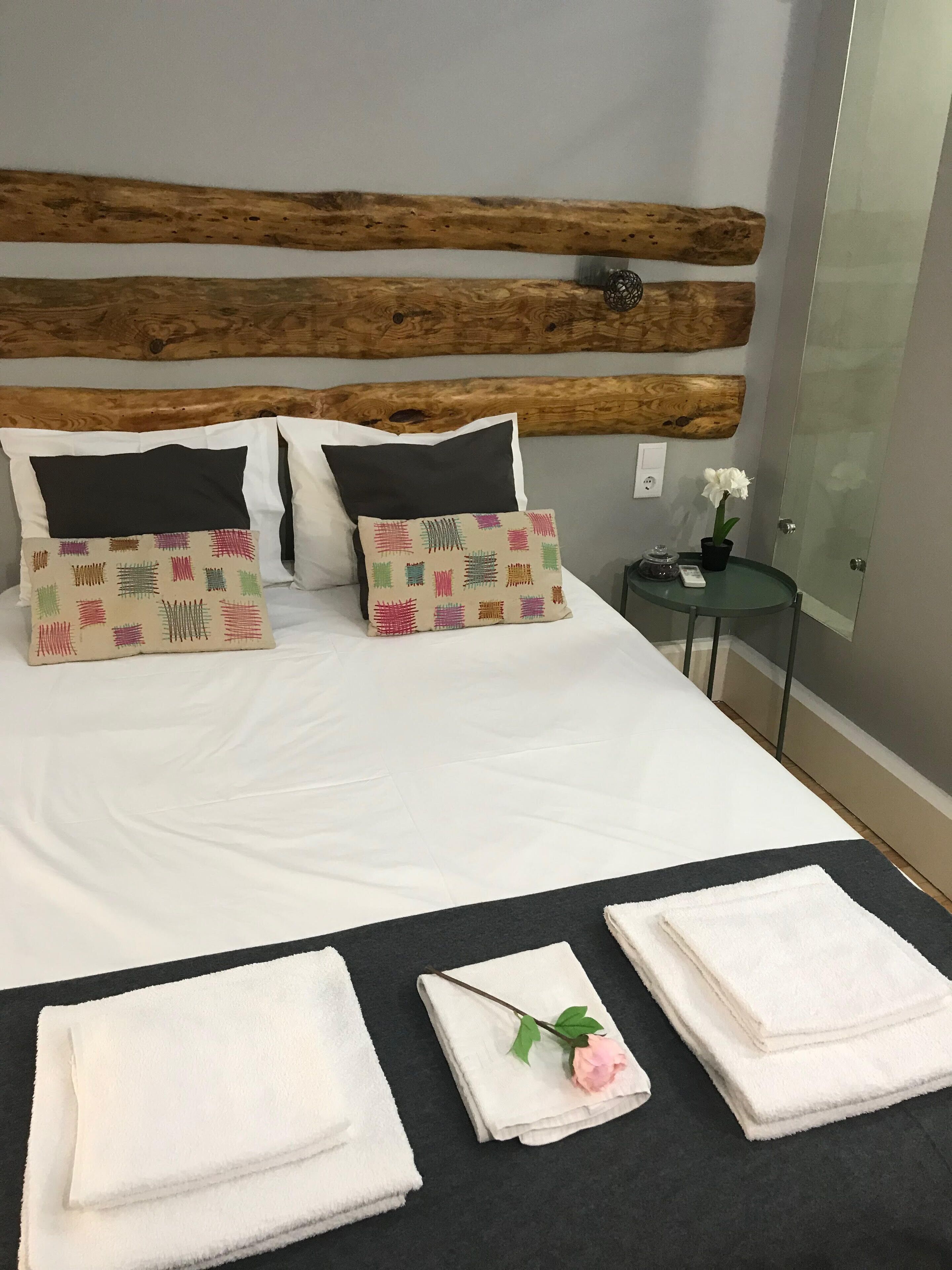 A neatly made bed with white linens, two black pillows, and colorful patterned pillows. There are folded white towels on a black table beside the bed, and a small potted plant with white flowers on a glass table.