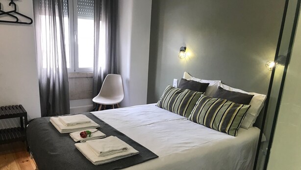 Una cama bien hecha con almohadas y toallas dobladas, una silla y una ventana con cortinas.