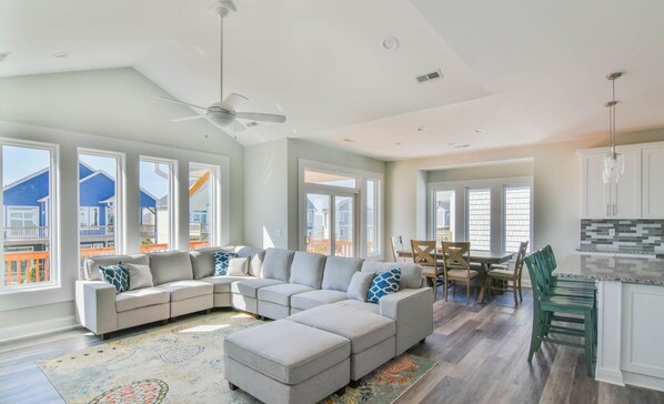 A spacious living room with a large L-shaped sectional sofa, a dining area with a wooden table and chairs, and a kitchen with white cabinets and a green bench.