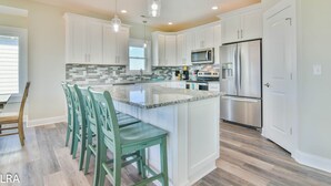 A modern kitchen with white cabinets, a stainless steel refrigerator, and a central island with bar stools. - Beach Bliss & Pool Days Await! (Surf City)