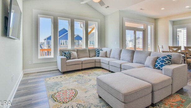 A spacious living room with a large L-shaped sectional sofa, a ceiling fan, and large windows offering views of neighboring houses. - Beach Bliss & Pool Days Await! (Surf City)
