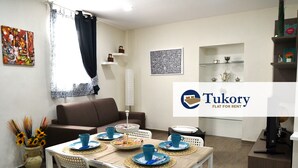 A modern living room with a dining area, featuring a sofa, a coffee table, and a dining table set with blue plates and cups. - Tukory - Flat For Rent - City Center Palermo (Palermo)