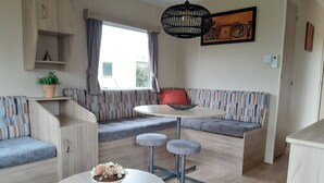 A modern living room with a sofa, a dining table with stools, a hanging light fixture, and a wall-mounted shelf with a plant. - RBR 244 - Beach Resort Kamperland (Kamperland)