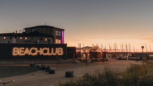 Un club de playa con un letrero de neón, un edificio con varias ventanas y una zona de muelles con barcos.