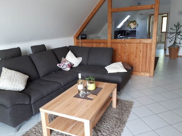 Ein modernes Wohnzimmer mit einem dunkelgrauen Ecksofa, einem Holztisch und einem durch eine Glastür einsehbarer Küchenbereich.