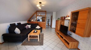Ein Wohnzimmer mit einem schwarzen Sofa, einem Holztisch, einem Fernseher auf einem Holzgestell und einer Treppe, die zu einer oberen Ebene führt.