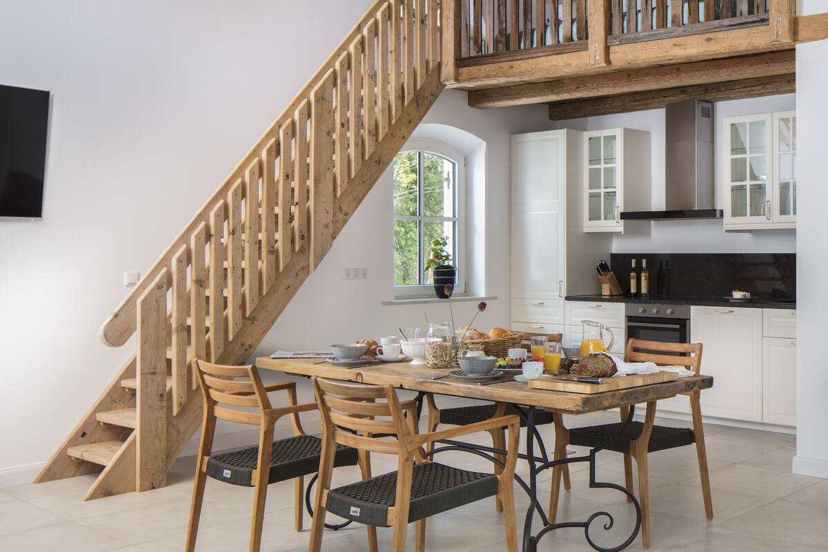 A dining area with a wooden staircase leading to a loft. There is a kitchen with white cabinets and a black countertop.