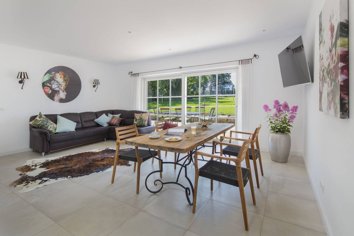 A spacious living room with a large window, a dining table with chairs, a sofa with cushions, and a cowhide rug.