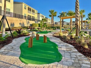 A landscaped area with a green artificial turf, palm trees, and a building with balconies.