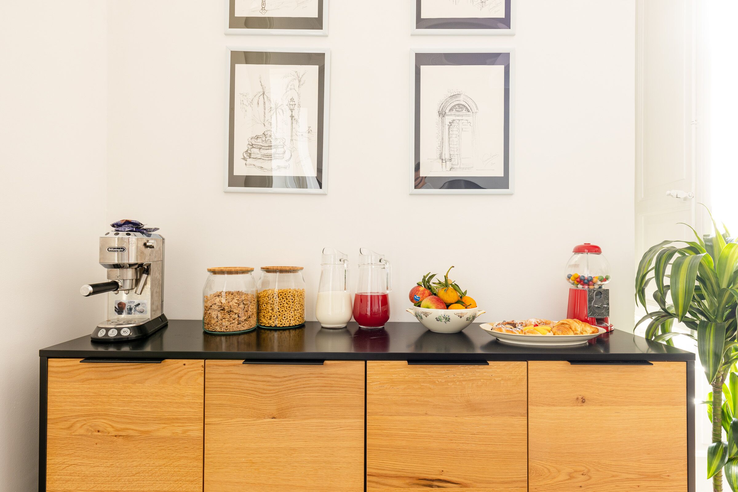 Una encimera de cocina con una cafetera, frascos de cereales, una jarra de leche, un bol de fruta y un recipiente rojo.