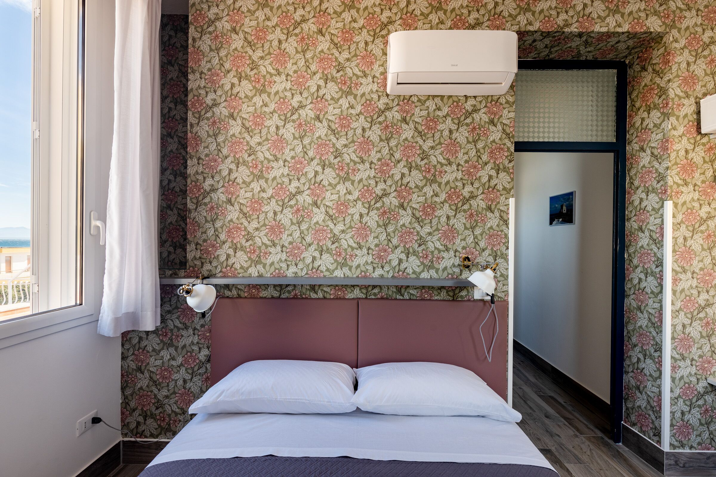 Une chambre à coucher avec un papier peint à motifs floraux, un lit recouvert de draps blancs, une table de chevet et une fenêtre avec un rideau.