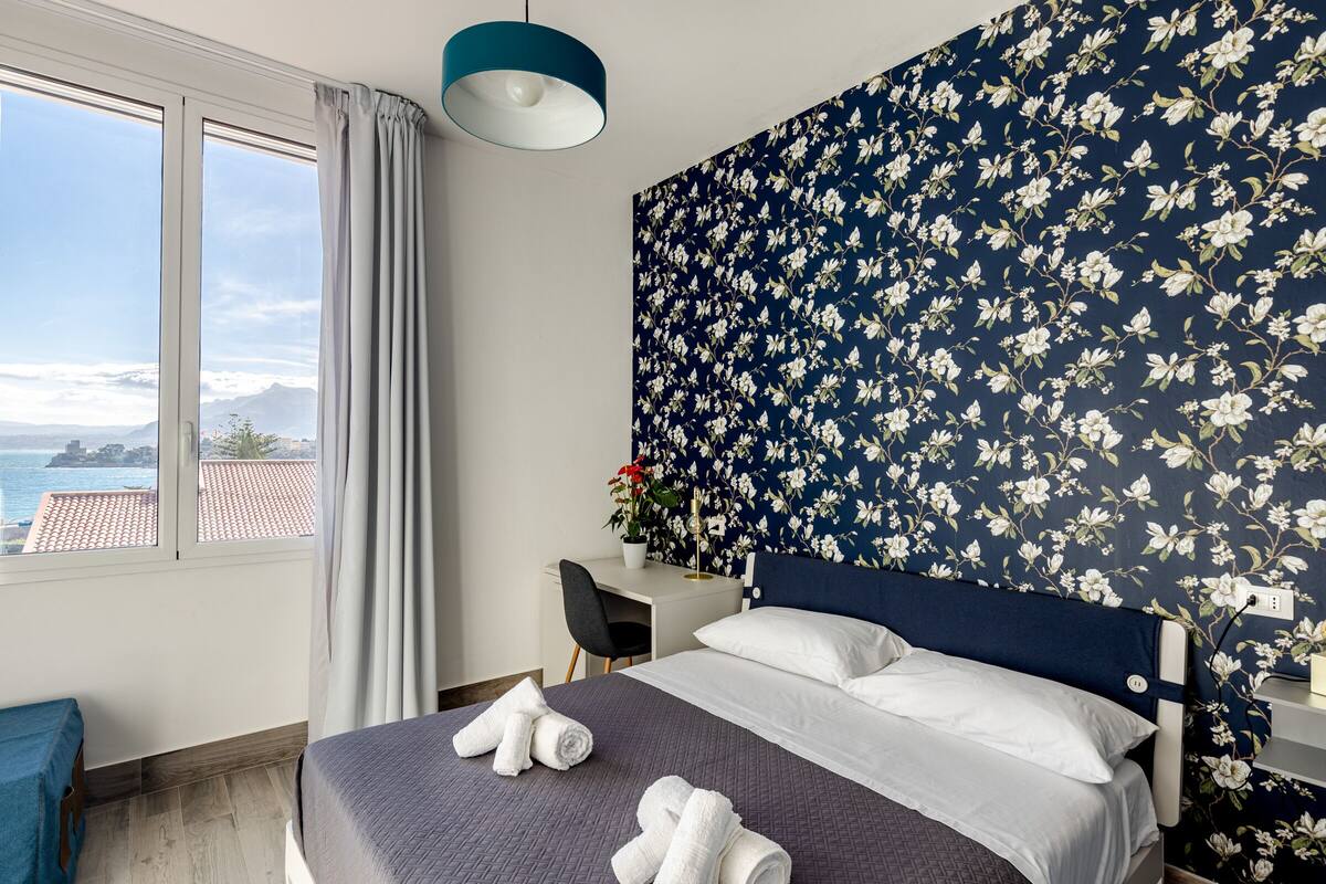 A hotel room with a bed, a desk, and a view of the sea.
