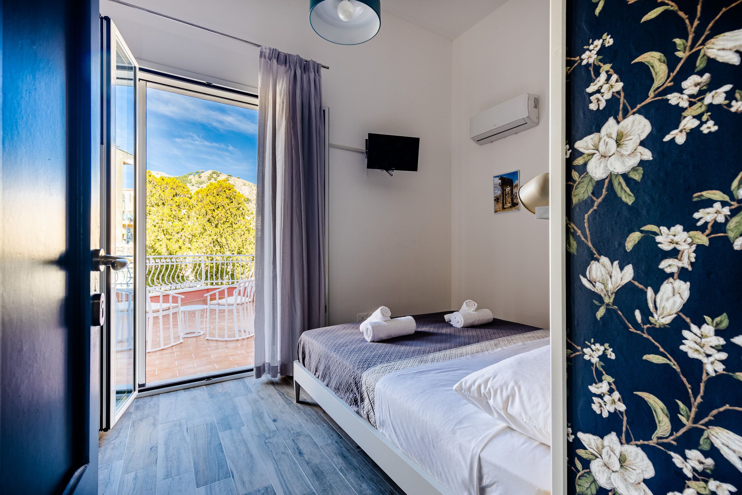 Un dormitorio con cama, televisor y balcón con vistas a árboles.