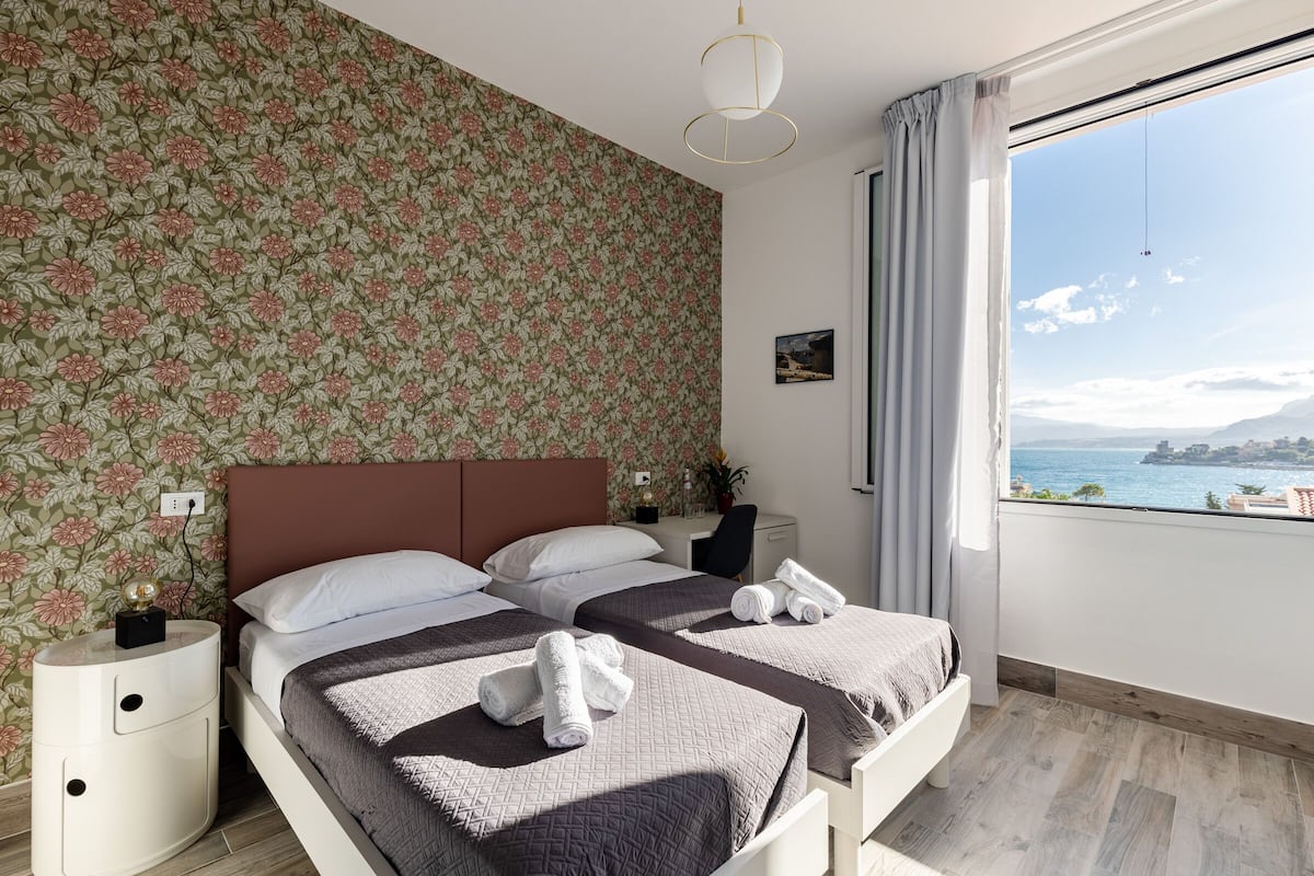 A bedroom with two beds, floral wallpaper, a view of the sea, and a nightstand with a lamp.