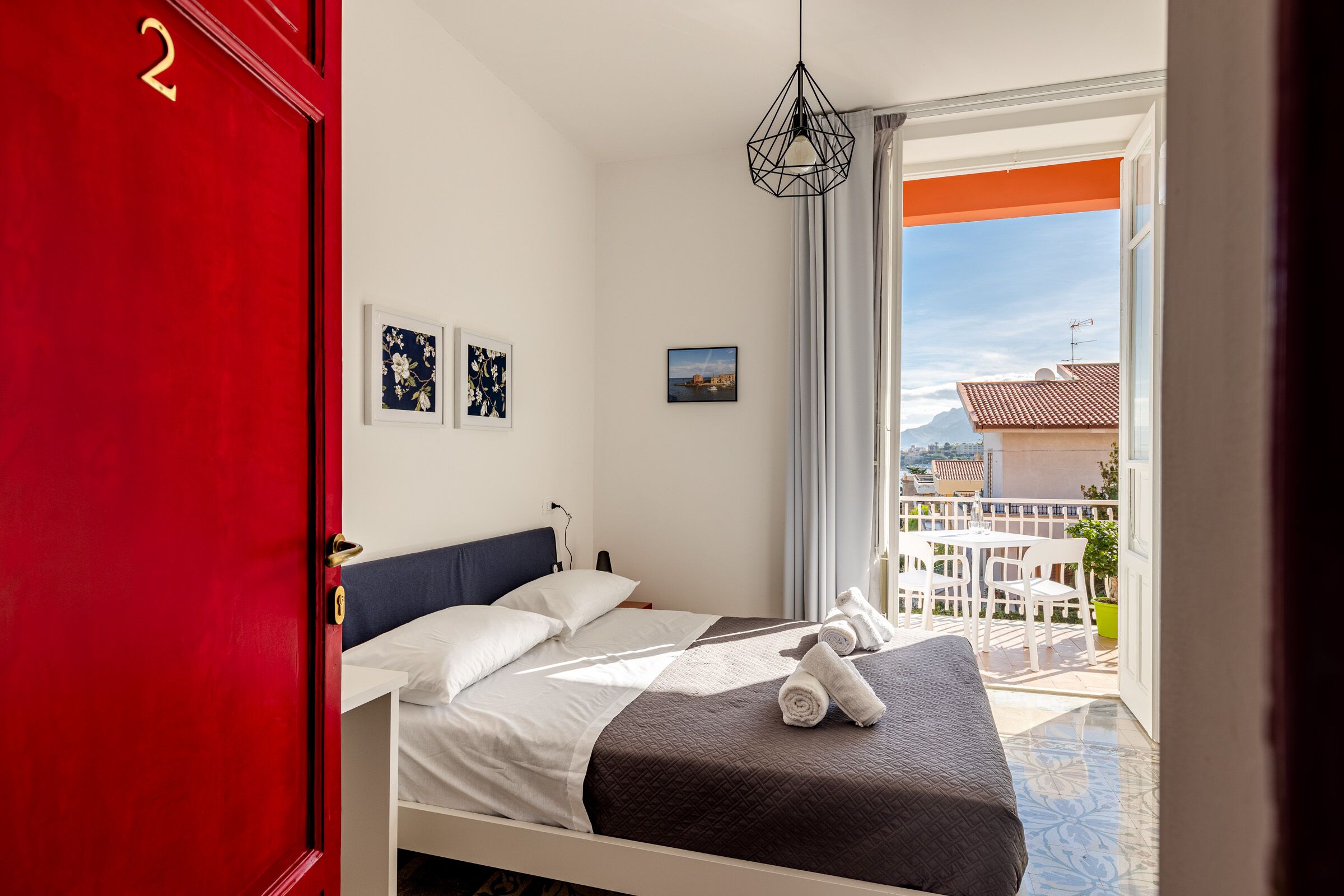 Hotelkamer met een bed, een rode deur met het nummer 2, een raam met uitzicht op gebouwen en een balkon met tuinmeubilair.