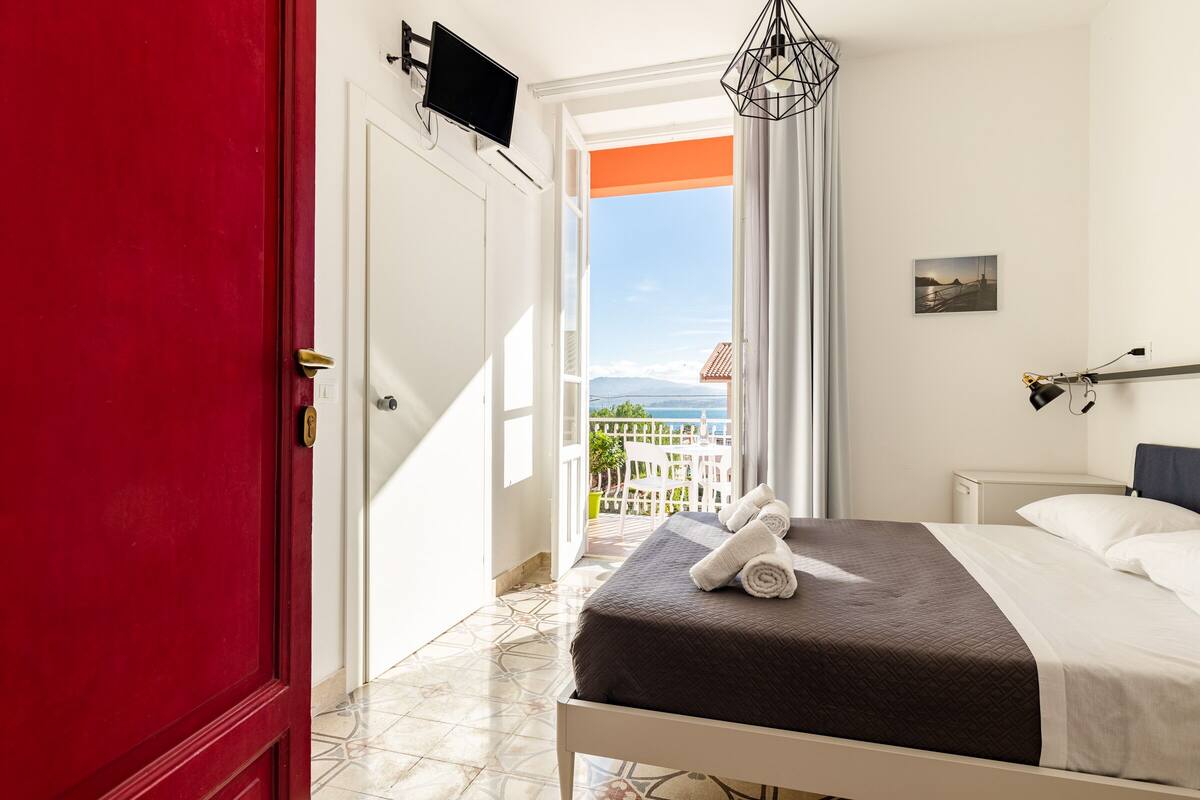 A hotel room with a bed, a TV, a view of the sea, and a balcony.