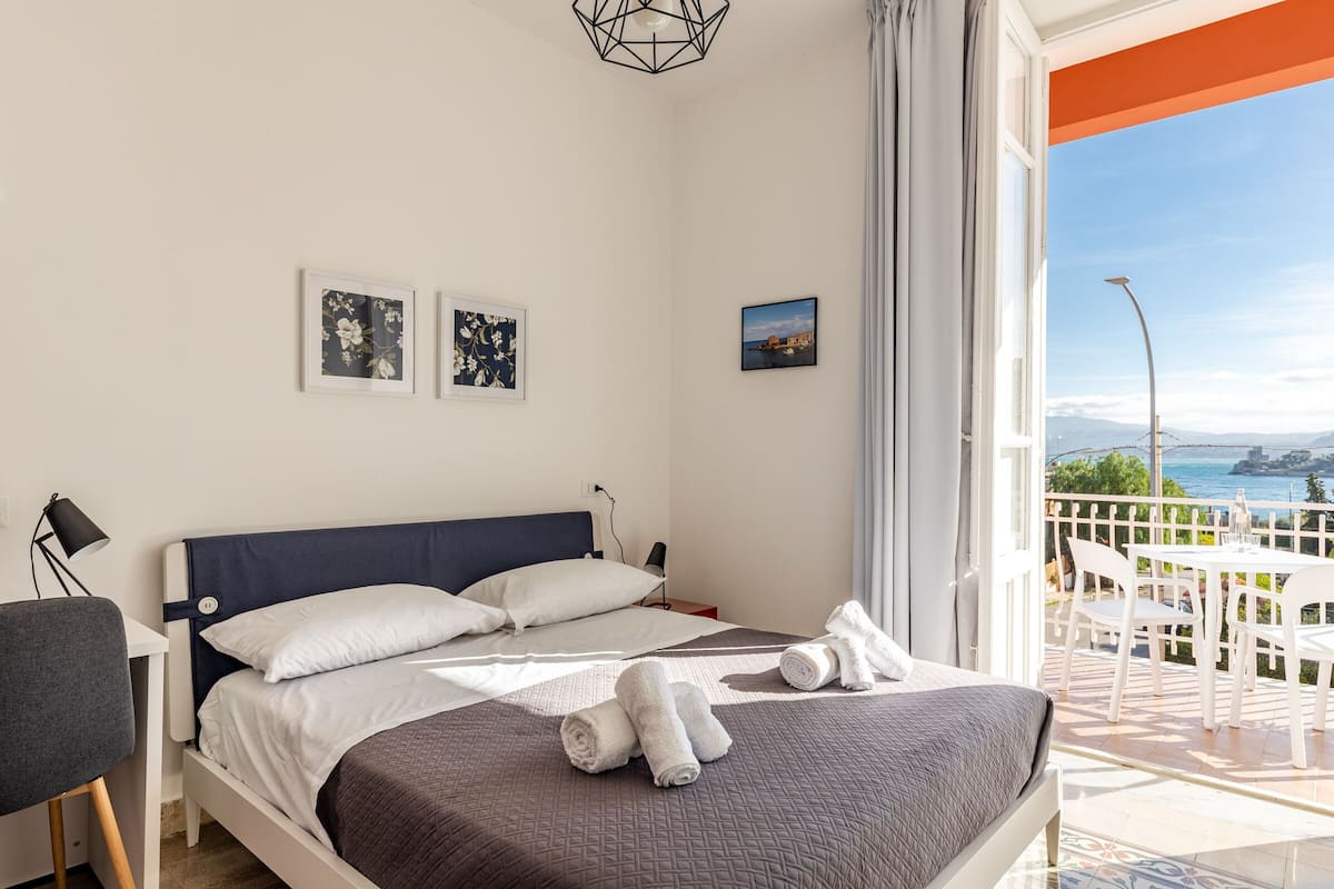 A neatly made bed with white and brown bedding, a desk with a chair, and a balcony with a view of the sea.