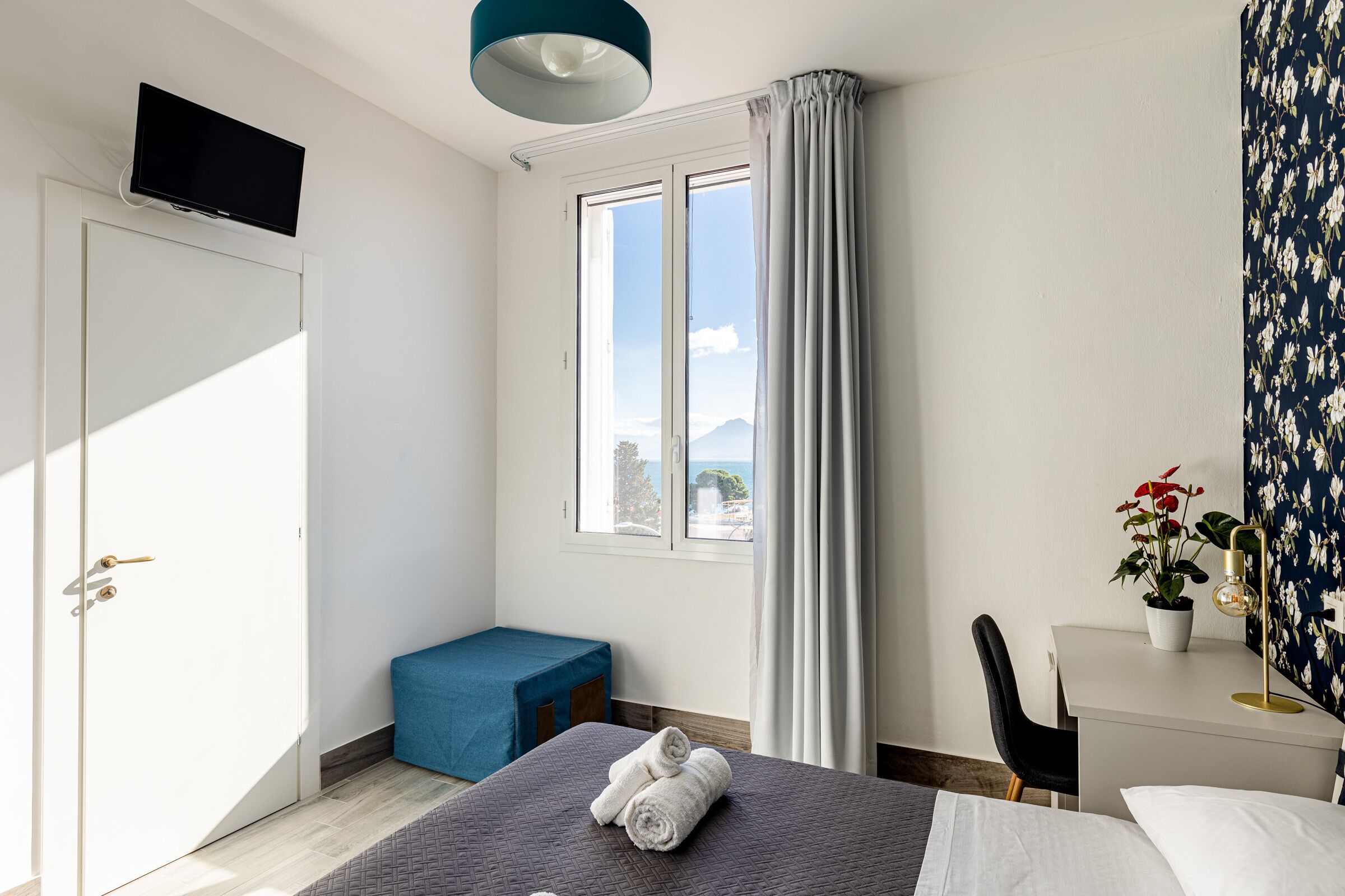 A bedroom with a bed, a desk, a chair, a TV, and a window with curtains.
